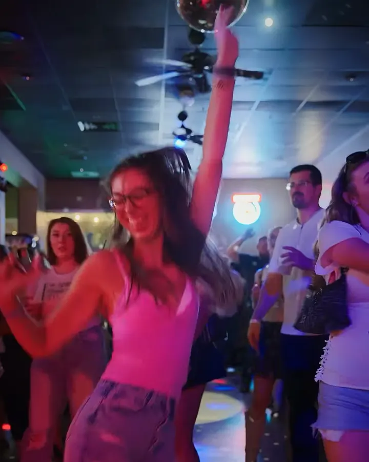 A woman dances with one arm raised under a disco ball on a crowded floor