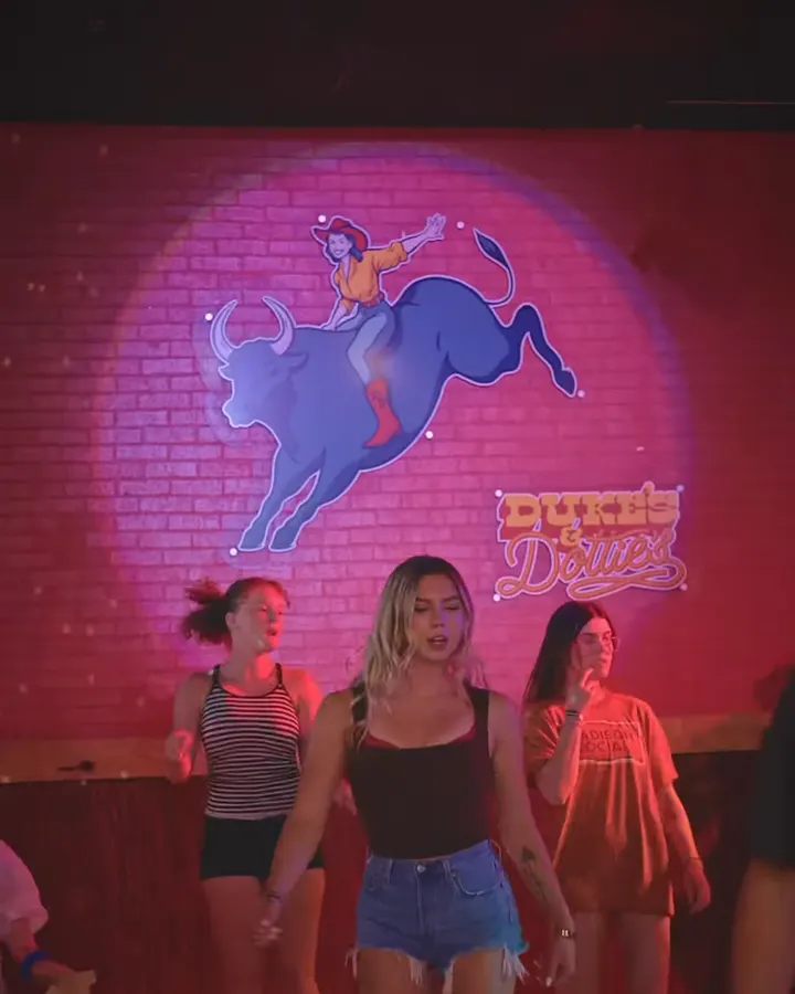 Dottie, the cowgirl riding the blue bull, lit up on the pink brick wall above three dancers