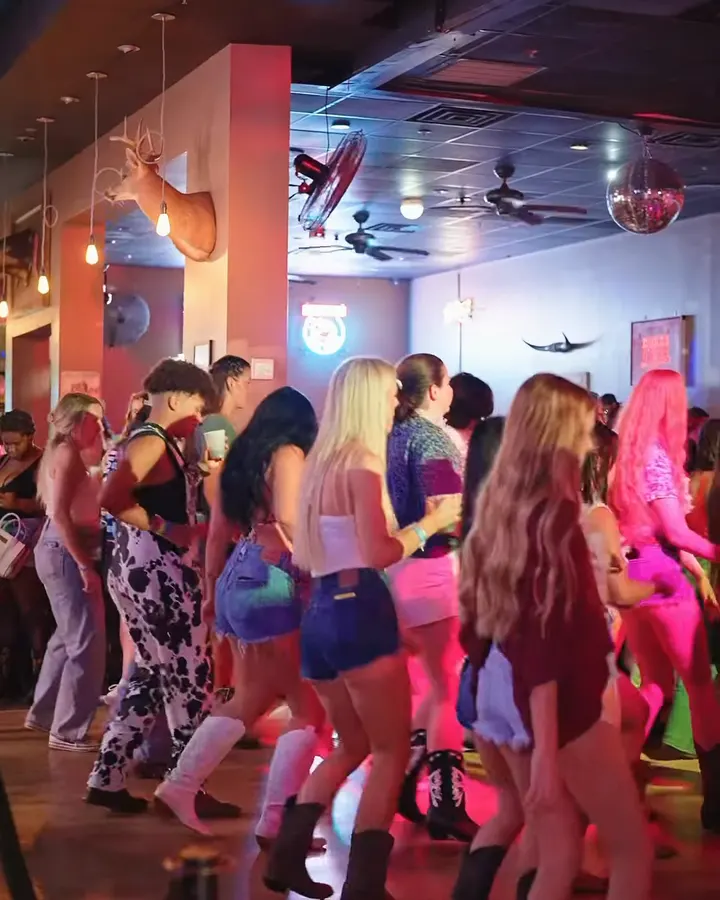 A line of dancers in boots on the wood floor, with a deer mount, a disco ball, and ceiling fans overhead